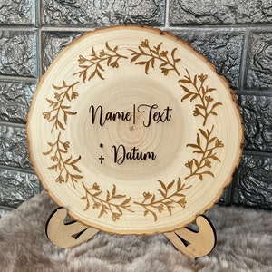 Op de afbeelding: Een ronde houten schijf met een decoratief, gegraveerd bloemmotief rond de rand. In het midden van de schijf staat de tekst "Name | Text * + Datum" gegraveerd in het hout.