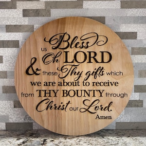 May include: A round wooden sign with the text "Bless us Oh LORD & these Thy gifts which we are about to receive from THY BOUNTY through Christ our Lord. Amen" in black script. The sign has a natural wood grain.