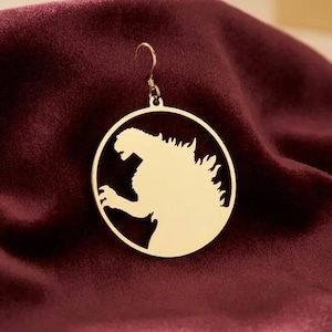 K&ouml;nnte beinhalten: Ein goldfarbener Ohrring mit einem kreisf&ouml;rmigen Design, das eine Silhouette eines br&uuml;llenden Monsters zeigt. Der Ohrring h&auml;ngt an einem kleinen Haken. Der Ohrring ist auf einem tiefroten, samtigen Stoff platziert.