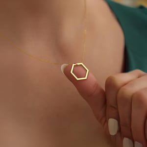 K&ouml;nnte beinhalten: Eine zarte goldene Halskette mit einem geometrischen sechseckigen Anh&auml;nger. Der Anh&auml;nger h&auml;ngt an einer feinen Kette. Die Halskette wird von einem Finger gehalten, wodurch das Design und die Farbe des Schmucks hervorgehoben werden.