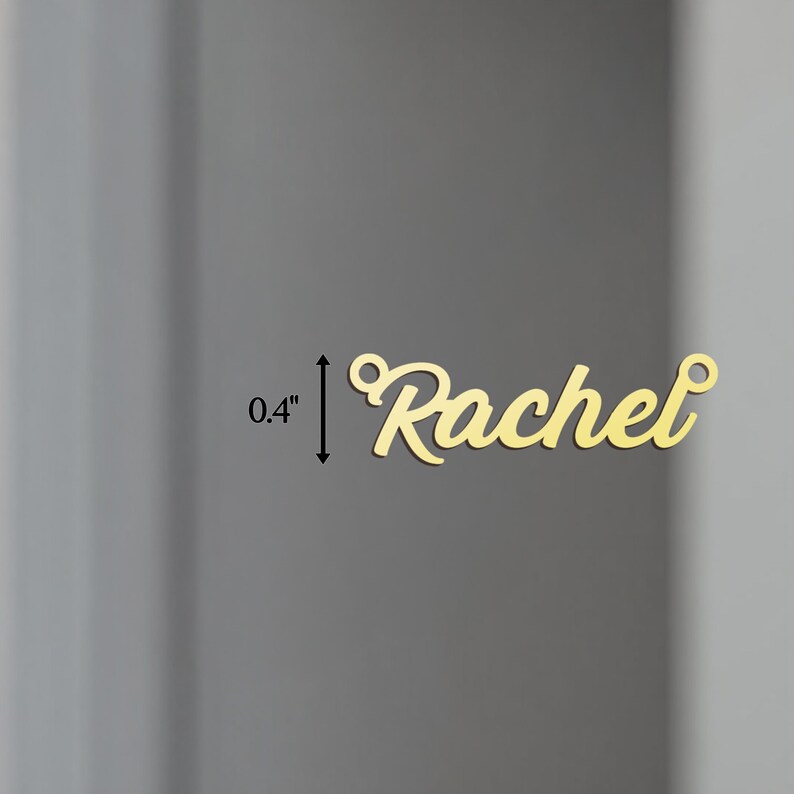 May include: Gold-colored metal charm with the name "Rachel" in a cursive font. The charm is 0.4 inches tall.