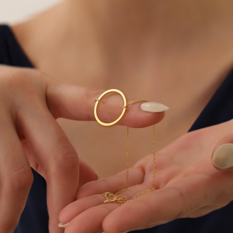 May include: A gold necklace with an open circle pendant. The pendant hangs from a fine chain. The necklace is held up by a person's fingers, highlighting the jewelry's design. The chain and pendant are a bright gold colour.