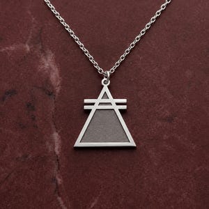 May include: A silver-toned necklace featuring a triangular pendant with a flat top and two horizontal bars crossing the upper section. The pendant has a smooth, matte-textured gray center framed by a polished silver border and hangs from a chain.