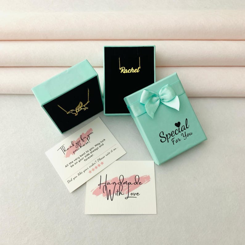 K&ouml;nnte beinhalten: Draufsicht auf Schmuckgeschenkboxen und Karten. Zwei goldene Halsketten werden in offenen Boxen pr&auml;sentiert. Eine Box ist schwarz, die andere t&uuml;rkis. Auf den Karten steht "Vielen Dank f&uuml;r Ihren Einkauf" und "Handmade With Love."