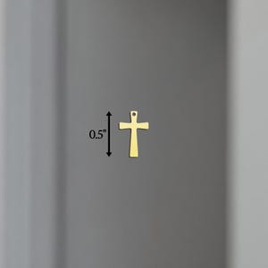 Puede incluir: Un peque&ntilde;o colgante con forma de cruz de color dorado, de aproximadamente 1,3 cm de altura, se muestra sobre un fondo gris neutro. La cruz tiene un dise&ntilde;o simple y cl&aacute;sico con un peque&ntilde;o lazo en la parte superior para colgar.