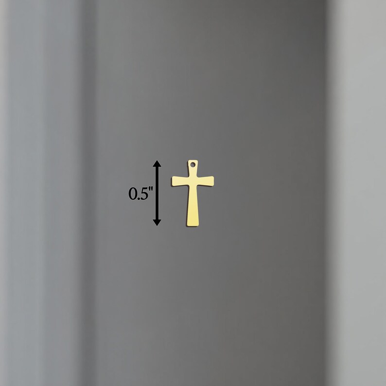 Puede incluir: Un peque&ntilde;o colgante con forma de cruz de color dorado, de aproximadamente 1,3 cm de altura, se muestra sobre un fondo gris neutro. La cruz tiene un dise&ntilde;o simple y cl&aacute;sico con un peque&ntilde;o lazo en la parte superior para colgar.