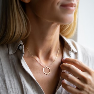 K&ouml;nnte beinhalten: Eine goldfarbene Halskette mit einem geometrischen sechseckigen Anh&auml;nger. Der Anh&auml;nger h&auml;ngt an einer zarten Kette. Die Halskette wird &uuml;ber einem hellfarbigen Hemd getragen.