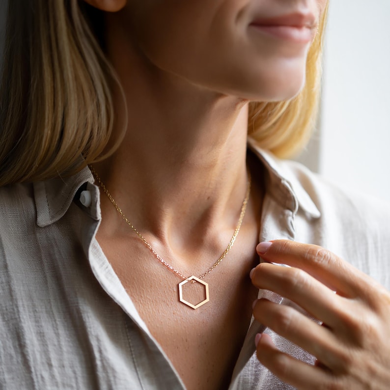 K&ouml;nnte beinhalten: Eine goldfarbene Halskette mit einem geometrischen sechseckigen Anh&auml;nger. Der Anh&auml;nger h&auml;ngt an einer zarten Kette. Die Halskette wird &uuml;ber einem hellfarbigen Hemd getragen.
