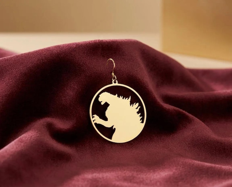 K&ouml;nnte beinhalten: Ein goldfarbener Ohrring mit einem kreisf&ouml;rmigen Design, das eine Silhouette eines br&uuml;llenden Monsters zeigt. Der Ohrring h&auml;ngt an einem kleinen Haken. Der Ohrring ist auf einem tiefroten, samtigen Stoff platziert.