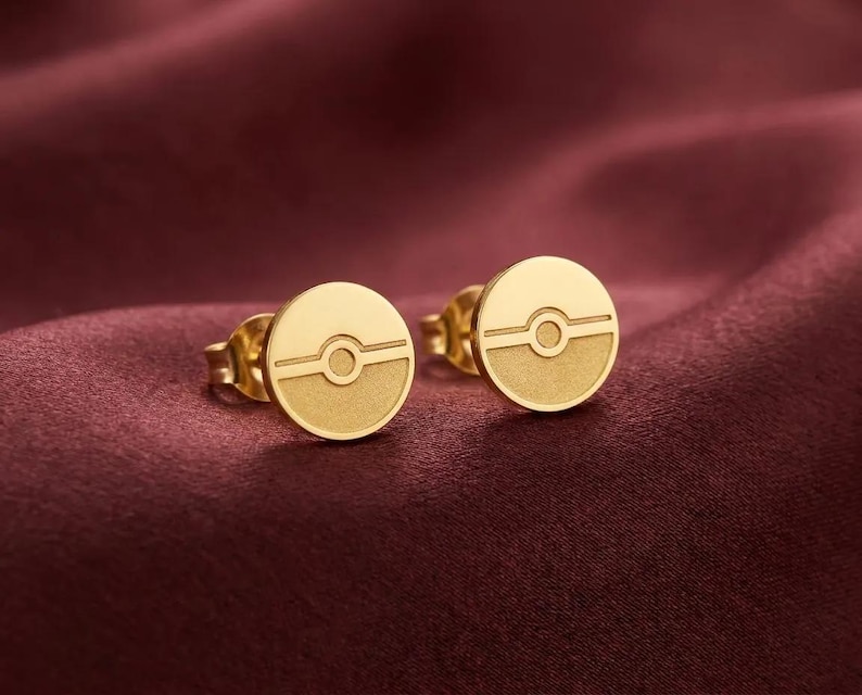 Peut inclure: Une paire de boucles d'oreilles &agrave; clous dor&eacute;es pr&eacute;sentant un motif circulaire avec une ligne horizontale et un cercle central, rappelant une sph&egrave;re. Les boucles ont une moiti&eacute; sup&eacute;rieure lisse et une moiti&eacute; inf&eacute;rieure textur&eacute;e et mate.