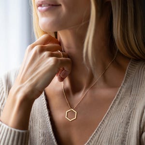 K&ouml;nnte beinhalten: Eine goldfarbene Halskette mit einem sechseckigen Anh&auml;nger. Der Anh&auml;nger ist eine einfache, geometrische Form, die an einer zarten Kette h&auml;ngt. Die Halskette wird &uuml;ber einem neutralfarbenen Pullover getragen.