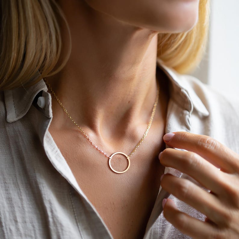 May include: A gold necklace with a fine chain and a circular pendant. The necklace is worn against a light-coloured shirt, creating a simple and elegant look. The pendant is a perfect circle.