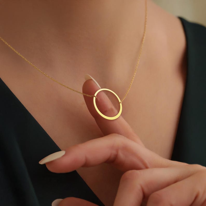 May include: A gold necklace with a fine chain and a circular pendant. The pendant is a simple, open circle design. The necklace is shown against a dark background.
