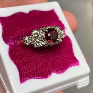 May include: A silver ring with a large, round, deep red gemstone at the center, flanked by smaller, clear stones. The ring is set in a white box with a pink velvet interior, showcasing the jewelry.