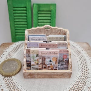 May include: A miniature wooden magazine rack filled with small magazines, with the text "Jeanne d'Arc Living" visible on the covers. The rack is light-colored, with a distressed finish. Two green shutters are in the background.