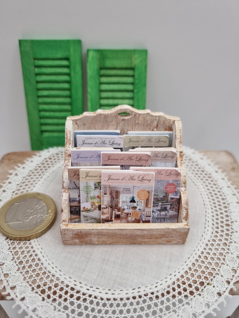 May include: A miniature wooden magazine rack filled with small magazines, with the text "Jeanne d'Arc Living" visible on the covers. The rack is light-colored, with a distressed finish. Two green shutters are in the background.