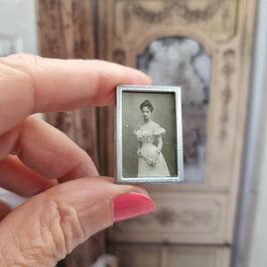 Puede incluir: Un marco plateado con una fotografía en blanco y negro de una mujer con un vestido blanco. La mujer está de pie y mirando a la cámara. El marco mide aproximadamente 5 cm de alto y 2,5 cm de ancho.