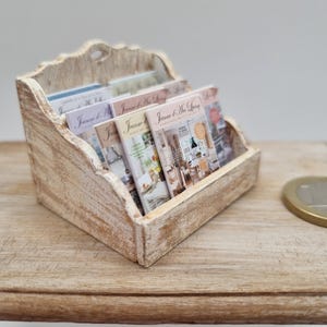 May include: A miniature wooden magazine rack with a distressed white finish, holding several small magazines. The magazines have colorful covers with the text "Jeanne d'Arc Living". The rack sits on a light-colored wooden surface.