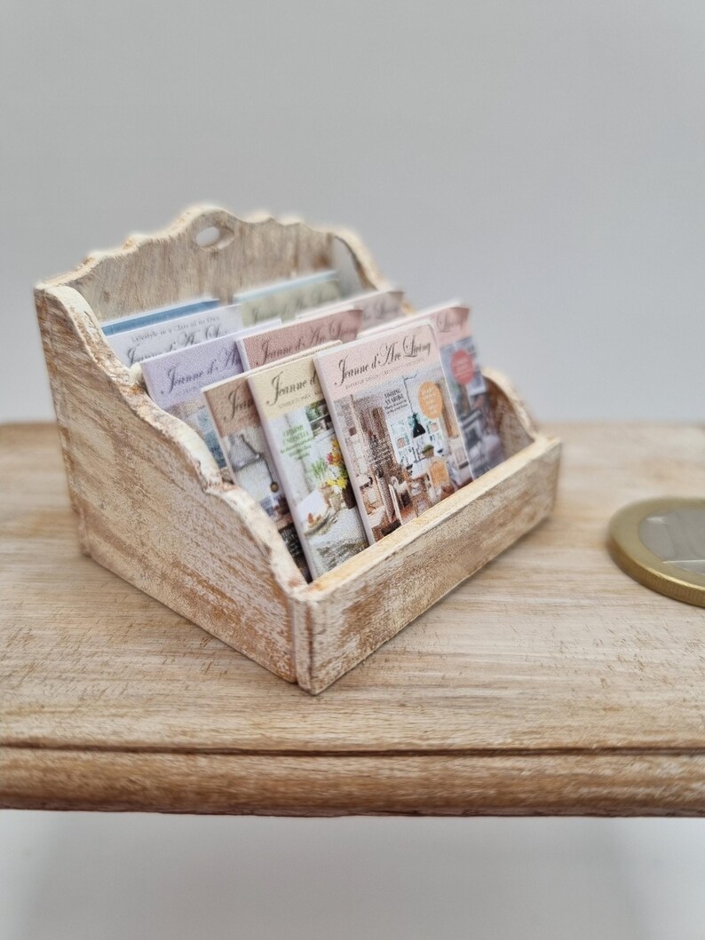 May include: A miniature wooden magazine rack with a distressed white finish, holding several small magazines. The magazines have colorful covers with the text "Jeanne d'Arc Living". The rack sits on a light-colored wooden surface.