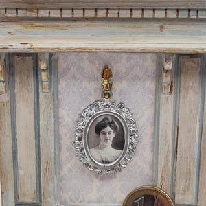 Op de afbeelding: Miniatuur portret van een vrouw in vintage stijl in een lijst. De ovale lijst is zilverkleurig en hangt aan een goudkleurige bevestiging. Het portret staat tegen een achtergrond van een lichtgekleurde muur met een bloemmotief. Een euro munt ligt op de voorgrond.