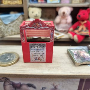 Könnte beinhalten: Ein rotes Miniatur-Puppentheater aus Holz mit einem Schild, auf dem Professor Fezzywigs Travelling Puppet Theatre steht, einem Wappen an der Oberseite und Abbildungen von Figuren, platziert auf einer Oberfläche neben einer Münze und einem kleinen Buch.