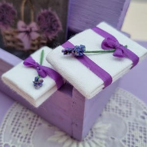 May include: Two small white fabric boxes with purple ribbon and lavender sprigs on top, sitting inside a purple wooden box.