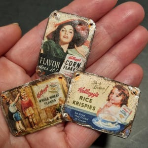 Op de afbeelding: Drie miniatuur vintage Kellogg's ontbijtgranen reclameborden. De borden tonen retro-advertenties voor Corn Flakes, Whole Wheat Flakes en Rice Krispies. Elk bord heeft een verweerde, verouderde uitstraling met zichtbare roest.