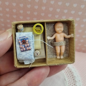 Op de afbeelding: Miniatuur knutseldoos voor poppenhuis met een kleine babypop, klosjes garen, stofresten en een klein pakketje met het woord "BIRANINIS" erop gedrukt. De doos wordt in een hand gehouden.