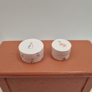 以下が含まれることがあります： 小さな丸い白い箱が2つ。それぞれにウサギと花のイラストが水彩で描かれています。箱は茶色の表面に置かれており、装飾品やコレクションアイテムのようです。