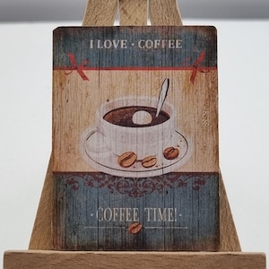 Puede incluir: Un pequeño caballete de madera exhibe un letrero rectangular con un diseño temático de café. El letrero presenta una taza de café, granos de café y las frases "I LOVE COFFEE" y "COFFEE TIME!" en un estilo vintage.