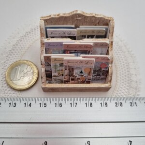 May include: Miniature wooden magazine rack filled with small magazines. The rack is a light brown color and has a decorative top. The magazines have colorful covers with text that reads "Jaume d'Art Living". A ruler shows the rack is approximately 7 inches wide.