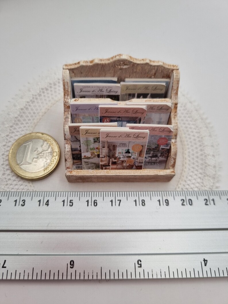 May include: Miniature wooden magazine rack filled with small magazines. The rack is a light brown color and has a decorative top. The magazines have colorful covers with text that reads "Jaume d'Art Living". A ruler shows the rack is approximately 7 inches wide.