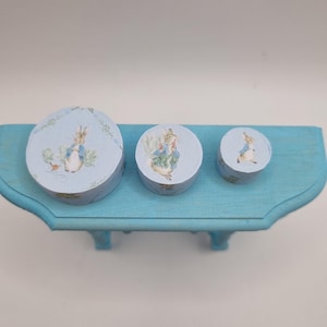 May include: Three round, light blue boxes of varying sizes, each adorned with a Peter Rabbit illustration. The boxes are arranged on a matching blue shelf, creating a cohesive and charming display. The boxes are perfect for storing small items.