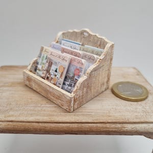 May include: A miniature wooden magazine rack with a distressed finish, holding several small magazines. The magazines have colorful covers with text. A coin rests on the wooden surface next to the rack.