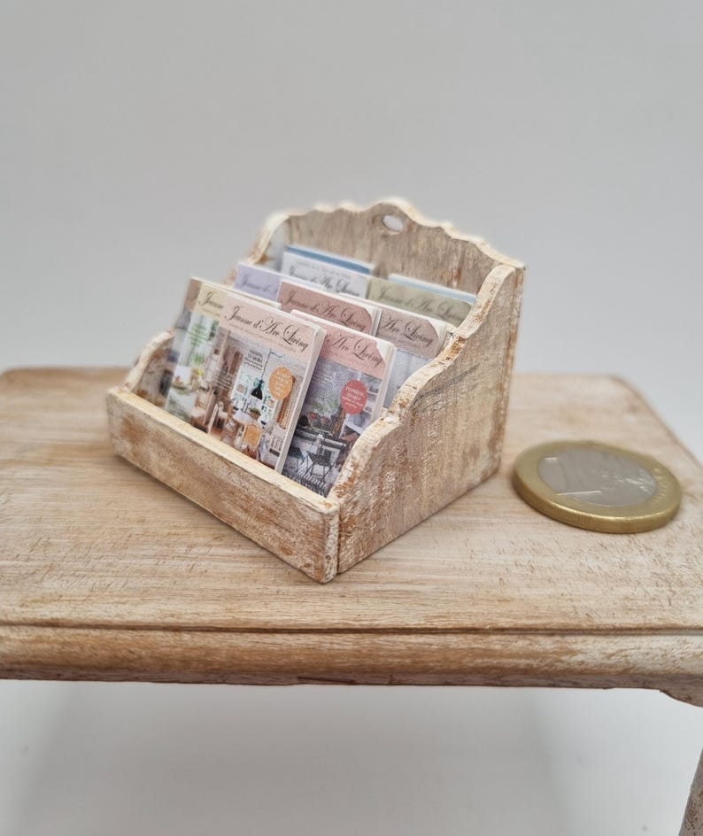 May include: A miniature wooden magazine rack with a distressed finish, holding several small magazines. The magazines have colorful covers with text. A coin rests on the wooden surface next to the rack.