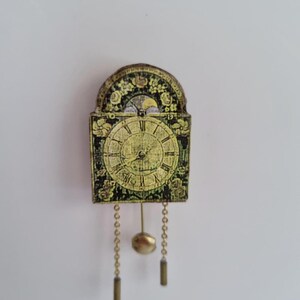 Puede incluir: Un pequeño reloj de estilo antiguo con una esfera cuadrada y una parte superior arqueada. El reloj presenta un diseño floral dorado y negro, un péndulo y dos pesas colgantes. La esfera del reloj tiene números romanos y manecillas ornamentadas.