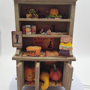 Könnte beinhalten: Ein hölzernes Miniatur-Küchenbuffet, dekoriert für Halloween, mit drei Regalen voller kleiner Kürbisse, Konservendosen, einem mit Happy beschrifteten Kuchen, einem Black Hat Society-Schild, einer Vogelscheuchenfigur und einem gerahmten Porträt.
