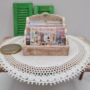 May include: A miniature wooden magazine rack filled with magazines titled "Jeanne d'Arc Living." The rack is light brown and sits on a small table with a white lace doily. Green shutters are in the background.