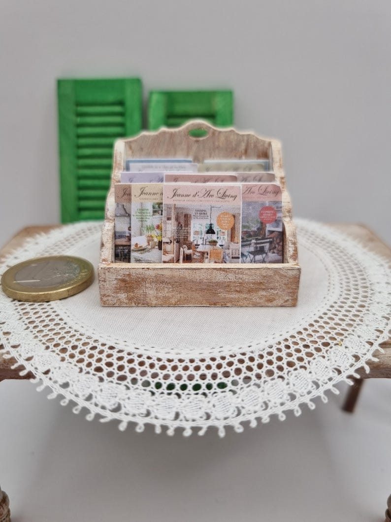 May include: A miniature wooden magazine rack filled with magazines titled "Jeanne d'Arc Living." The rack is light brown and sits on a small table with a white lace doily. Green shutters are in the background.