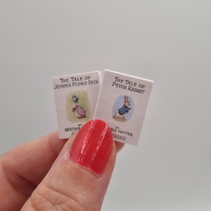 May include: Two miniature books, "The Tale of Jemima Puddle-Duck" and "The Tale of Peter Rabbit," are held in a hand. The book covers feature illustrations of the characters. The titles and author names are printed on the covers.