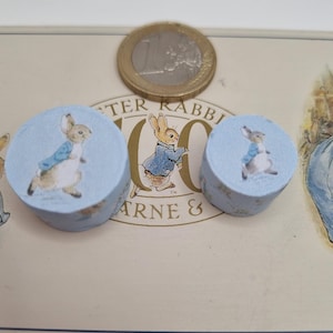 Puede incluir: Dos pequeños objetos cilíndricos de color azul claro con ilustraciones de Peter Rabbit. Una moneda de oro descansa sobre una lata vintage con las palabras "Peter Rabbit" y "Warne & Co" impresas. Probablemente artículos decorativos o de manualidades.