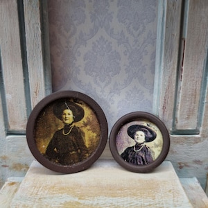Op de afbeelding: Twee antieke bruin omlijste portretten. De sepia-afbeeldingen tonen vrouwen met hoeden en kettingen. De ronde frames rusten op een verweerd, lichtgekleurd oppervlak. Achtergrond met bloemenbehang.