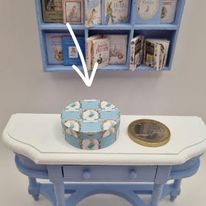 Op de afbeelding: Een kleine, ronde blauw-witte doos met een bloemmotief staat op een wit-blauwe tafel. Een euromunt ligt naast de doos. Op de achtergrond staat een plank met miniatuurboeken.