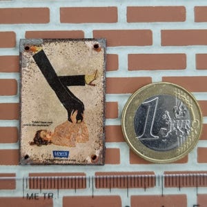 May include: A small, rectangular metal sign with a vintage advertisement featuring a woman and a man's legs. The sign has a distressed, aged appearance with the Levi's logo. A Euro coin and a ruler are next to the sign.