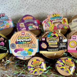 May include: A collection of round, tan-colored containers featuring various colorful labels for Mystery Squishy Dumpling toys. The packaging displays text including Squeezy Dumpling, Vos Squishy Bao, and Mystery Dumpling, with illustrations of small, round, colorful dumpling characters.