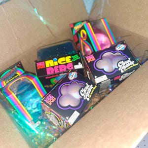 May include: A cardboard box containing several Nee Doh sensory toys in their original packaging. Visible items include two Cloud Pleaser boxes, a Nice Berg box, and an Urban Drop box, all featuring colorful rainbow accents and black branding.