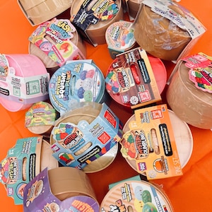 May include: A collection of round, plastic-encased Mystery Squishy Dumpling toys in various colors and packaging designs. Each container features colorful labels with text such as The Viral Mystery Dumpling, ages 3+, and various scent labels.
