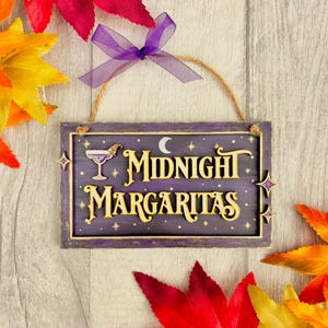 May include: A rectangular purple wooden sign with the text Midnight Margaritas in raised cream lettering. The sign features a small cocktail glass with a witch hat, a crescent moon, and stars. It hangs from a twine string with a purple ribbon.