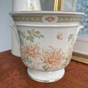 May include: A white ceramic planter with a floral design featuring orange and cream chrysanthemums and green leaves. The planter has a gold rim and handles. A small butterfly is also depicted.