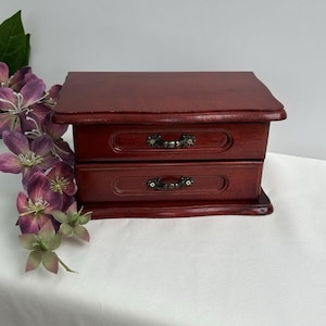 May include: A small, two-drawer jewelry box made of dark red wood. The box has ornate metal handles and sits on a white surface. Artificial purple flowers are arranged to the left of the box.
